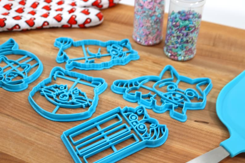 Puede incluir: Un juego de seis cortadores de galletas de pl&aacute;stico azul con forma de mapaches. Los cortadores est&aacute;n sobre una superficie de madera con chispas en frascos en el fondo.