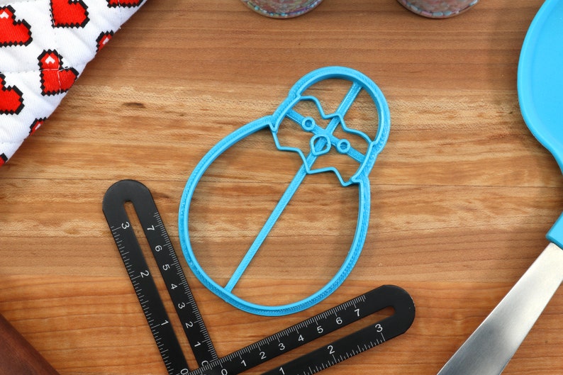 May include: A blue plastic cookie cutter shaped like a penguin. The cutter is on a wooden surface.