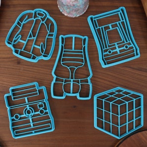 May include: A set of six blue plastic cookie cutters shaped like a jacket, roller skates, a video game, a camera, a Rubik's cube, and a cassette tape.