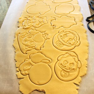 May include: A sheet of cookie dough with various cartoon character shapes cut out. The shapes include a mushroom, a star, a face with a crown, and a face with a mustache.