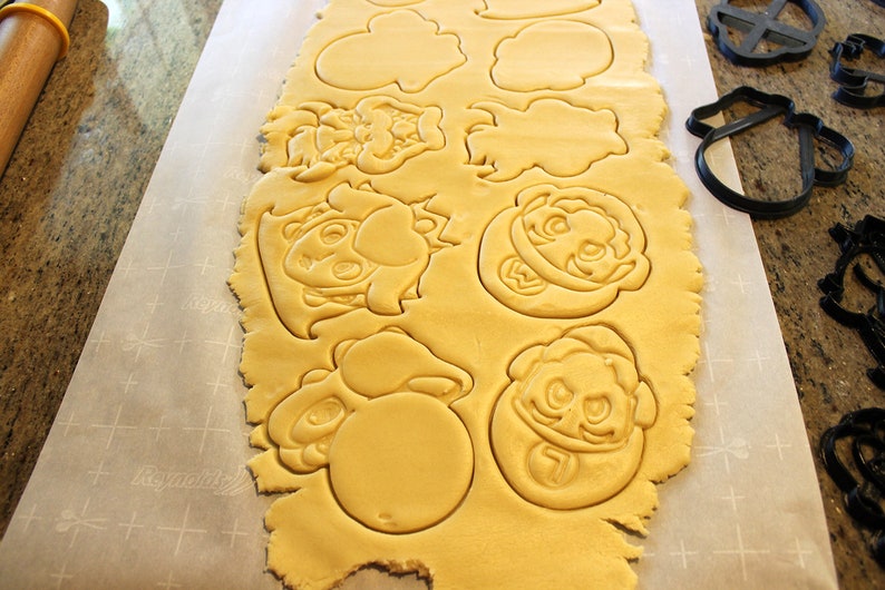 May include: A sheet of cookie dough with various cartoon character shapes cut out. The shapes include a mushroom, a star, a face with a crown, and a face with a mustache.