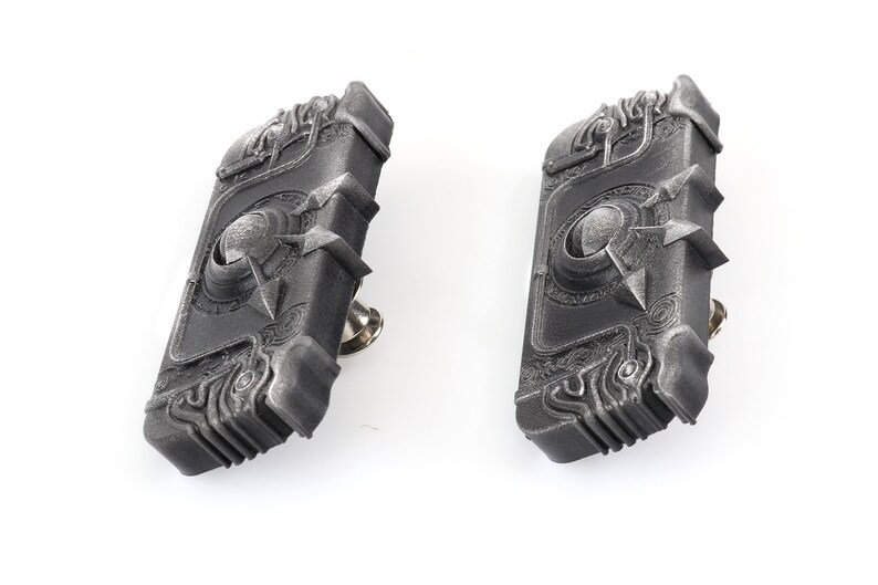 May include: Two silver-coloured metal book-shaped pins with intricate designs. The pins have a textured surface and a raised circular design in the centre.