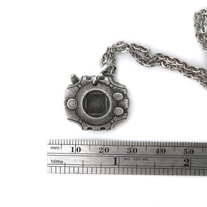 Puede incluir: Un collar plateado con un colgante con forma de dispositivo digital. El colgante tiene una pantalla cuadrada y una serie de botones alrededor del borde. El collar mide 60 mm de largo.