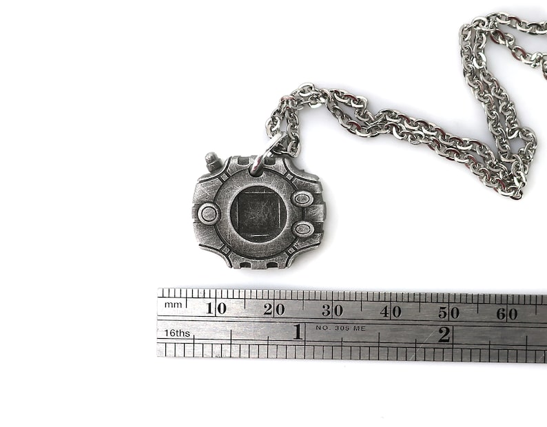 Puede incluir: Un collar plateado con un colgante con forma de dispositivo digital. El colgante tiene una pantalla cuadrada y una serie de botones alrededor del borde. El collar mide 60 mm de largo.