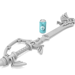 K&ouml;nnte beinhalten: Eine wei&szlig;e Plastik-Replik der Kingdom Key, ein schl&uuml;sself&ouml;rmiges Schwert aus der Kingdom Hearts-Videospielserie. Das Schwert liegt auf einer wei&szlig;en Oberfl&auml;che neben einer blauen Dose.