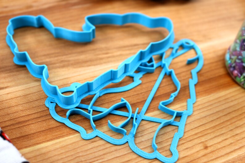 May include: Two blue plastic cookie cutters, one shaped like a heart and the other shaped like a person, are lying on a wooden surface.