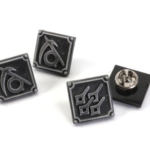 May include: Three silver metal pins with different designs. The first pin features a stylized symbol with a crescent moon and a circle. The second pin features a stylized symbol with a crescent moon and a circle. The third pin features a stylized symbol with four squares.