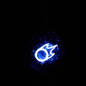 Puede incluir: Una bola de cristal azul con un s&iacute;mbolo de llama blanca brillante en su interior. El s&iacute;mbolo es un c&iacute;rculo con una llama que sale de la parte superior.
