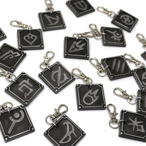 May include: A set of 16 silver keychains with different symbols on each. The keychains are black with silver accents and are all square shaped.