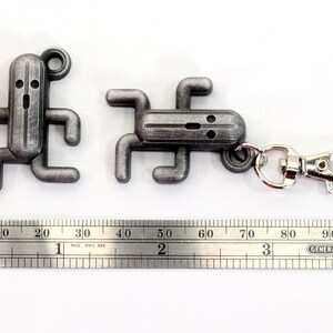 May include: Two silver metal keychain charms shaped like a video game character. The larger charm has a silver metal ring attached to it. A ruler is in the background.
