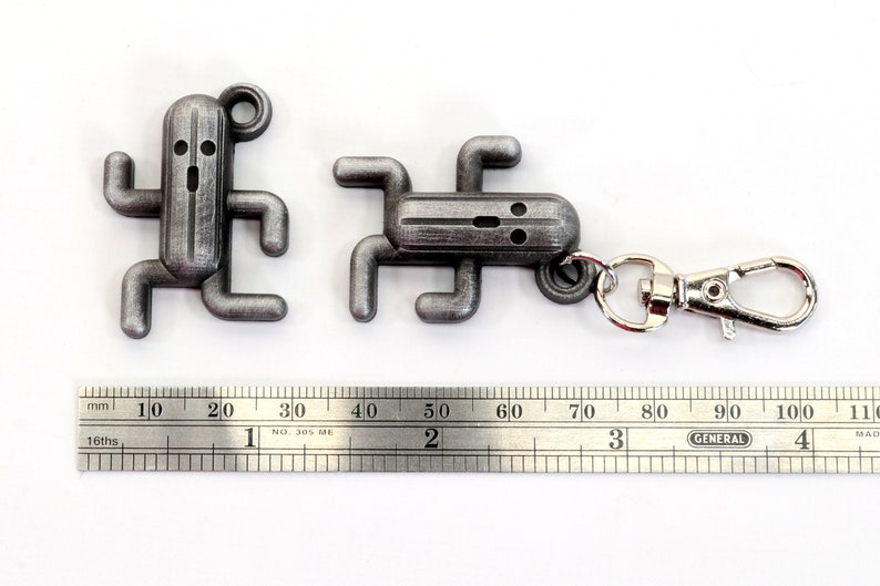 May include: Two silver metal keychain charms shaped like a video game character. The larger charm has a silver metal ring attached to it. A ruler is in the background.