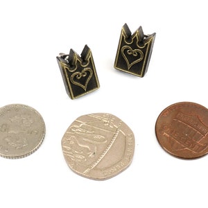 May include: Two small, dark metal earrings with a crown-like top and a heart design. The earrings are next to a penny, a British 50 pence coin, and a British 20 pence coin, all on a white background.