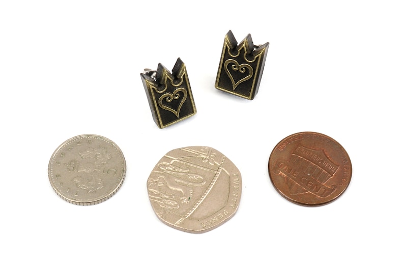 May include: Two small, dark metal earrings with a crown-like top and a heart design. The earrings are next to a penny, a British 50 pence coin, and a British 20 pence coin, all on a white background.