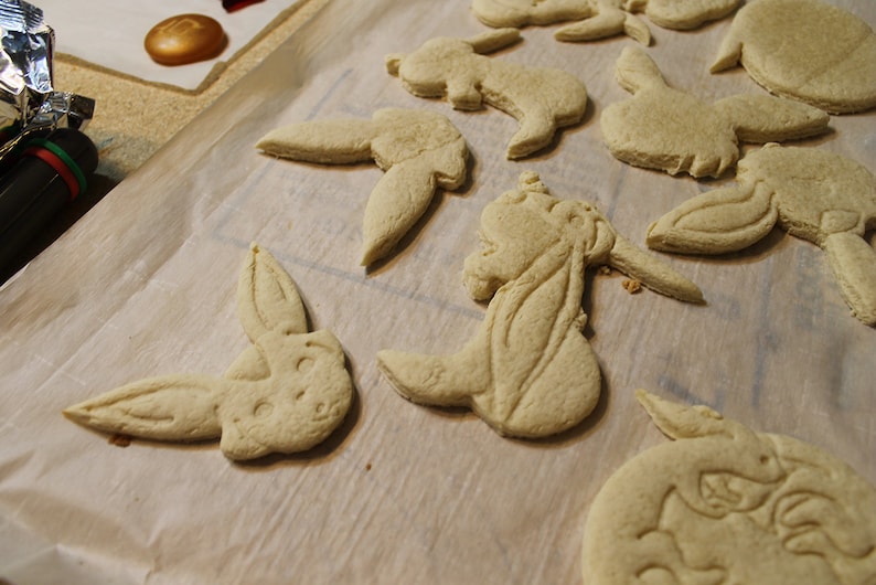 May include: A baking sheet lined with baking parchment holds a variety of unbaked, light brown sugar cookies. The cookies are shaped like bunnies and have detailed designs.