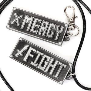 May include: A black cord necklace with two silver metal tags. The tags have pixelated text that reads "FIGHT" and "MERCY".