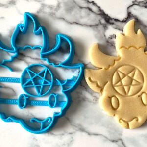 Pode incluir: Um cortador de biscoitos de pl&aacute;stico azul em forma de cabra de desenho animado com chifres e um pentagrama na testa. Ao lado dele est&aacute; um biscoito assado no mesmo formato.