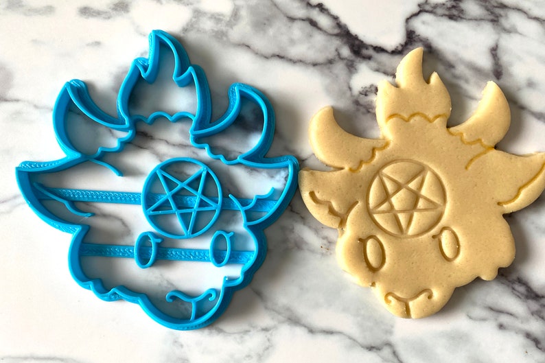Pode incluir: Um cortador de biscoitos de pl&aacute;stico azul em forma de cabra de desenho animado com chifres e um pentagrama na testa. Ao lado dele est&aacute; um biscoito assado no mesmo formato.