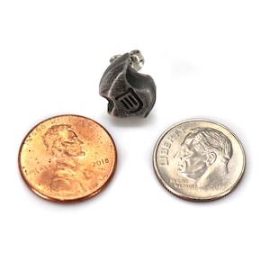May include: A small, textured, silver-toned metal charm with a stylized design and a small loop. A penny and a dime are next to the charm for size comparison. The charm has a dark, aged appearance.