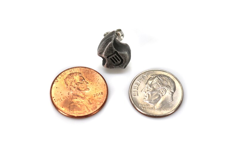 May include: A small, textured, silver-toned metal charm with a stylized design and a small loop. A penny and a dime are next to the charm for size comparison. The charm has a dark, aged appearance.