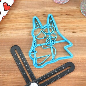 May include: A blue plastic cookie cutter shaped like a cartoon dog wearing sunglasses. The cutter is on a wooden surface.