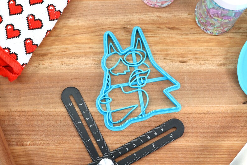 May include: A blue plastic cookie cutter shaped like a cartoon dog wearing sunglasses. The cutter is on a wooden surface.