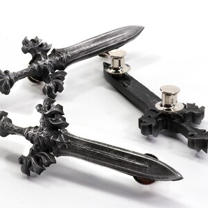 May include: Three silver-toned metal sword pins with intricate details. The swords have a pointed tip and a decorative hilt. The pins are attached to the back of the swords.