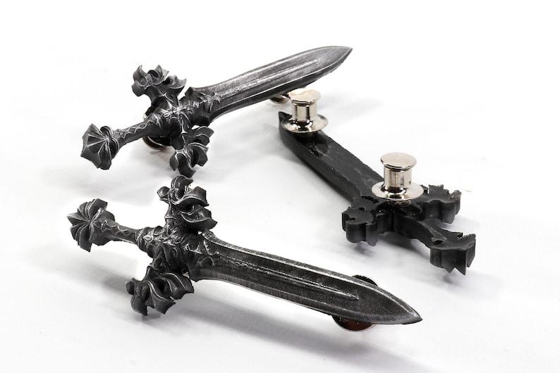 May include: Three silver-toned metal sword pins with intricate details. The swords have a pointed tip and a decorative hilt. The pins are attached to the back of the swords.