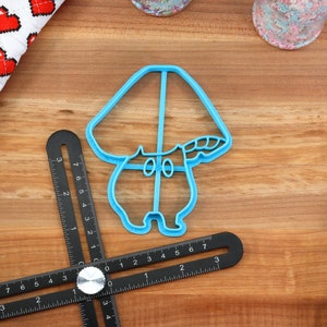 May include: A blue plastic cookie cutter shaped like a mushroom with a white stem and a brown cap. The cookie cutter is on a wooden surface.
