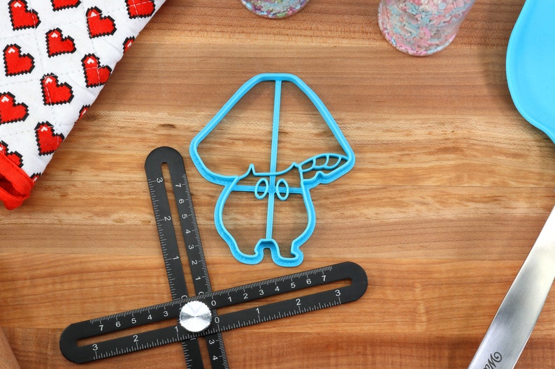 May include: A blue plastic cookie cutter shaped like a mushroom with a white stem and a brown cap. The cookie cutter is on a wooden surface.