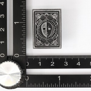 Op de afbeelding: Een zilverkleurige metalen pin met een zwart-wit gezichtontwerp. De pin is omgeven door een decoratieve rand en heeft een getextureerd oppervlak.