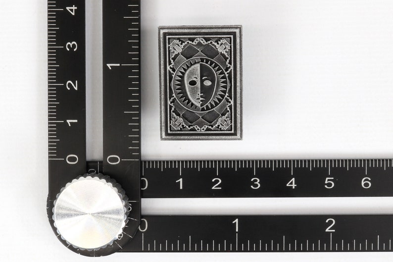 Op de afbeelding: Een zilverkleurige metalen pin met een zwart-wit gezichtontwerp. De pin is omgeven door een decoratieve rand en heeft een getextureerd oppervlak.