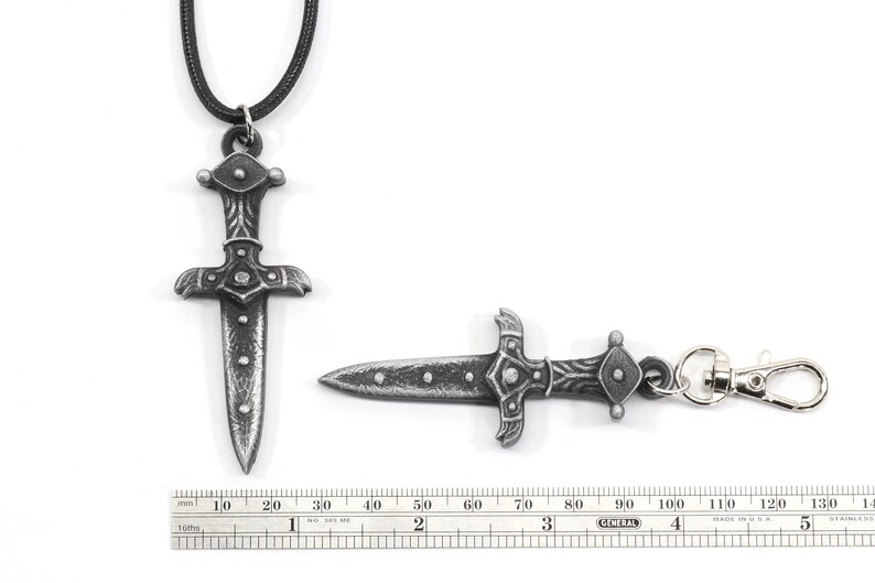 May include: A silver-toned dagger pendant with a black cord necklace and a matching silver-toned dagger keychain. The dagger has a detailed design with a pointed tip and a rounded handle. The pendant is approximately 7 cm long and the keychain is approximately 6 cm long.