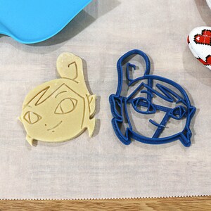 May include: Two cookie cutters, one blue and one beige, shaped like cartoon faces. The blue cutter is on a white surface.