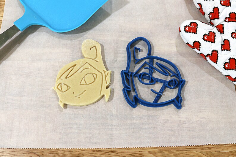 May include: Two cookie cutters, one blue and one beige, shaped like cartoon faces. The blue cutter is on a white surface.