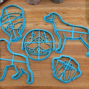 May include: Five blue plastic cookie cutters shaped like different dog breeds. The cutters are on a wooden surface.