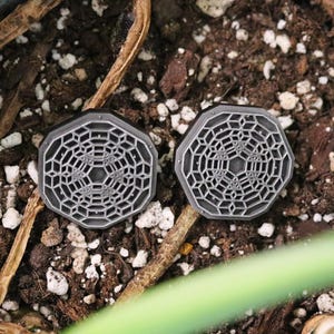 May include: Two gray octagonal-shaped earrings with a geometric design. The earrings are on a brown surface with white pebbles.