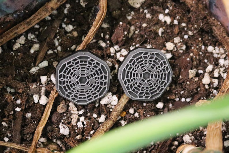 May include: Two gray octagonal-shaped earrings with a geometric design. The earrings are on a brown surface with white pebbles.