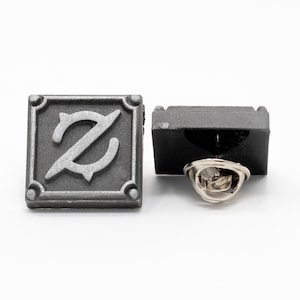 Op de afbeelding: Een zilverkleurige metalen pin met een vierkant ontwerp met een gestileerde letter 'Z' in het midden. De pin heeft een zwarte achterkant en een zilverkleurige pin achterkant.