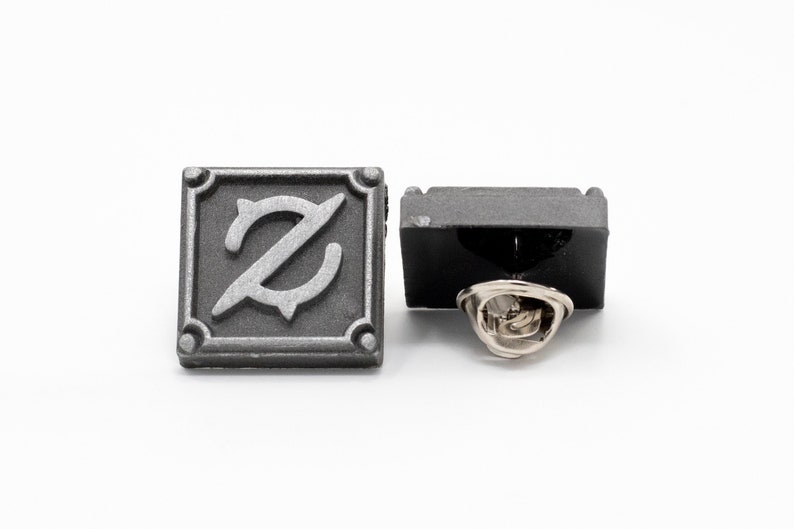 Op de afbeelding: Een zilverkleurige metalen pin met een vierkant ontwerp met een gestileerde letter 'Z' in het midden. De pin heeft een zwarte achterkant en een zilverkleurige pin achterkant.