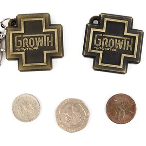 May include: Two gold-colored metal keychains with the word "GROWTH" on them. The keychains are in the shape of a cross. There are three coins below the keychains, two silver and one copper.