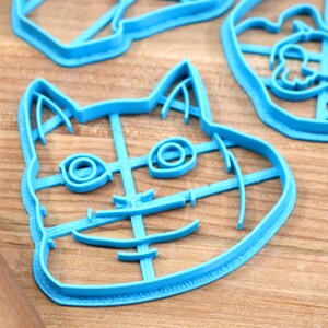 May include: A blue plastic cookie cutter shaped like a cat's face. The cutter has a detailed design with lines for the eyes, nose, and mouth.