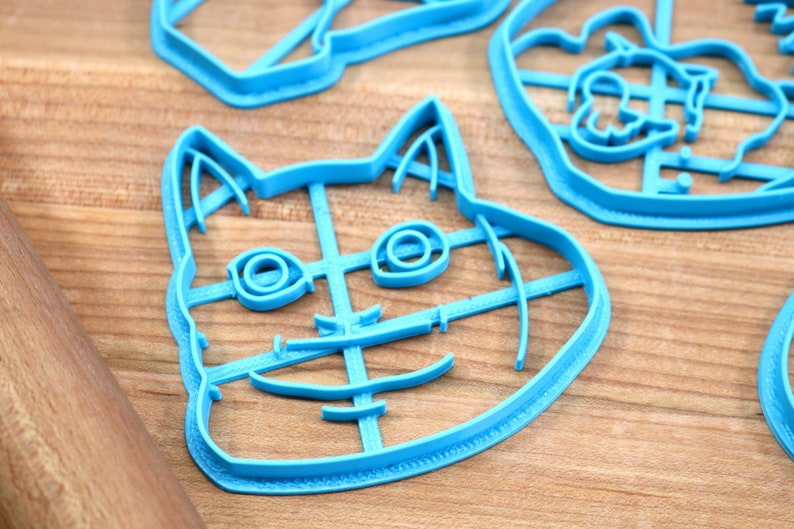 May include: A blue plastic cookie cutter shaped like a cat's face. The cutter has a detailed design with lines for the eyes, nose, and mouth.