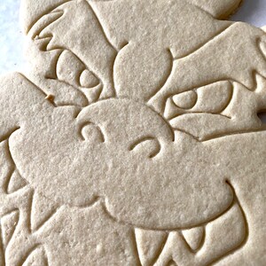 Op de afbeelding: Een close-up van een eenvoudige, ongebakken suikerkoek in de vorm van een drakenkop. De koek heeft een gedetailleerd ontwerp met ogen, een mond en tanden.