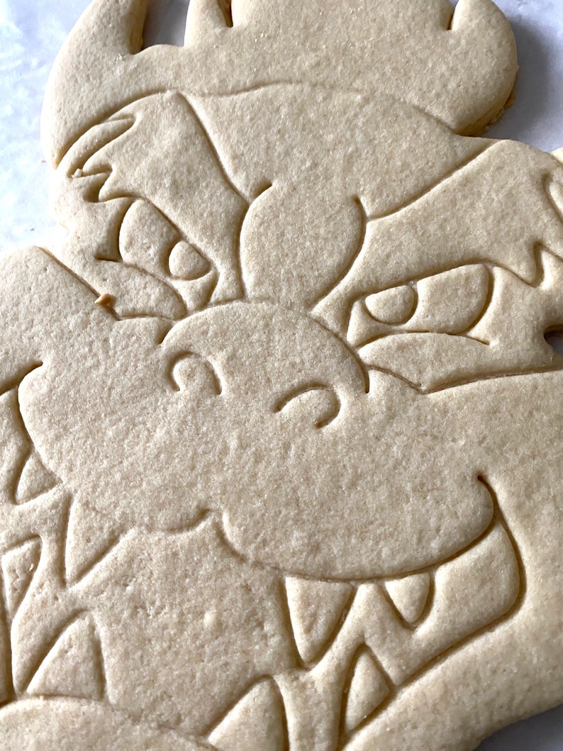 Op de afbeelding: Een close-up van een eenvoudige, ongebakken suikerkoek in de vorm van een drakenkop. De koek heeft een gedetailleerd ontwerp met ogen, een mond en tanden.