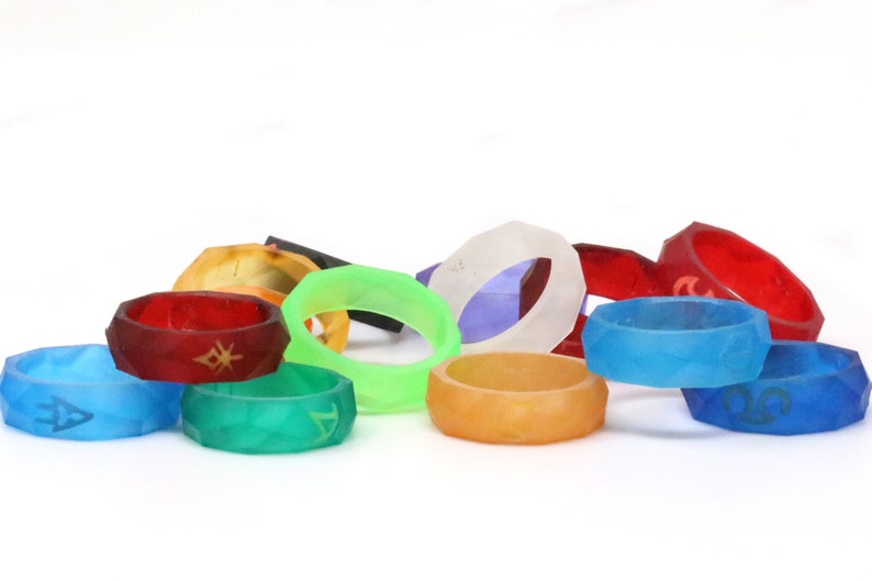 May include: A collection of colorful, geometric rings with different symbols and designs. The rings are made of a translucent material and are stacked on a white surface.