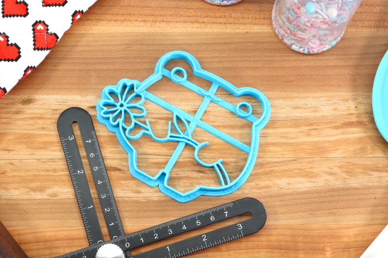 May include: A blue plastic cookie cutter shaped like a frog holding a flower. The cutter is on a wooden surface.
