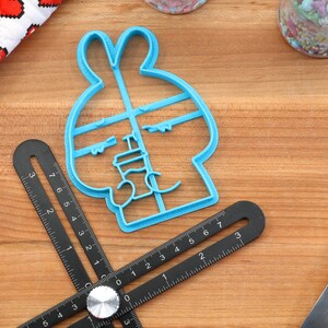 May include: A blue plastic cookie cutter shaped like a bunny rabbit with a face and ears. The cutter is on a wooden surface with a black ruler and a silver rolling pin.