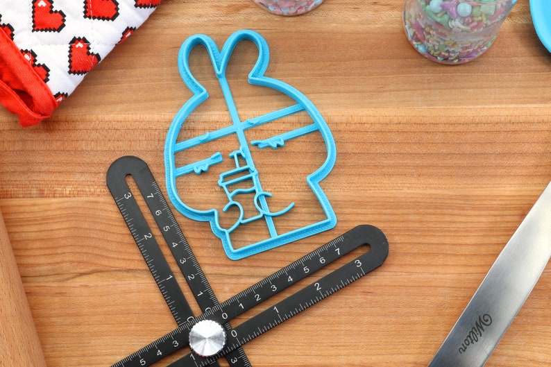 May include: A blue plastic cookie cutter shaped like a bunny rabbit with a face and ears. The cutter is on a wooden surface with a black ruler and a silver rolling pin.