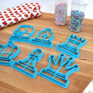 May include: A set of seven blue plastic cookie cutters shaped like chess pieces. The cutters are arranged on a wooden surface.