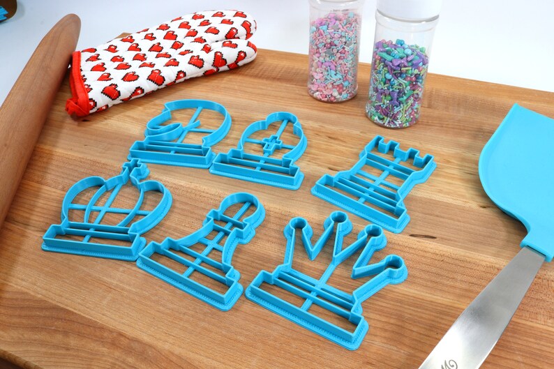 May include: A set of seven blue plastic cookie cutters shaped like chess pieces. The cutters are arranged on a wooden surface.